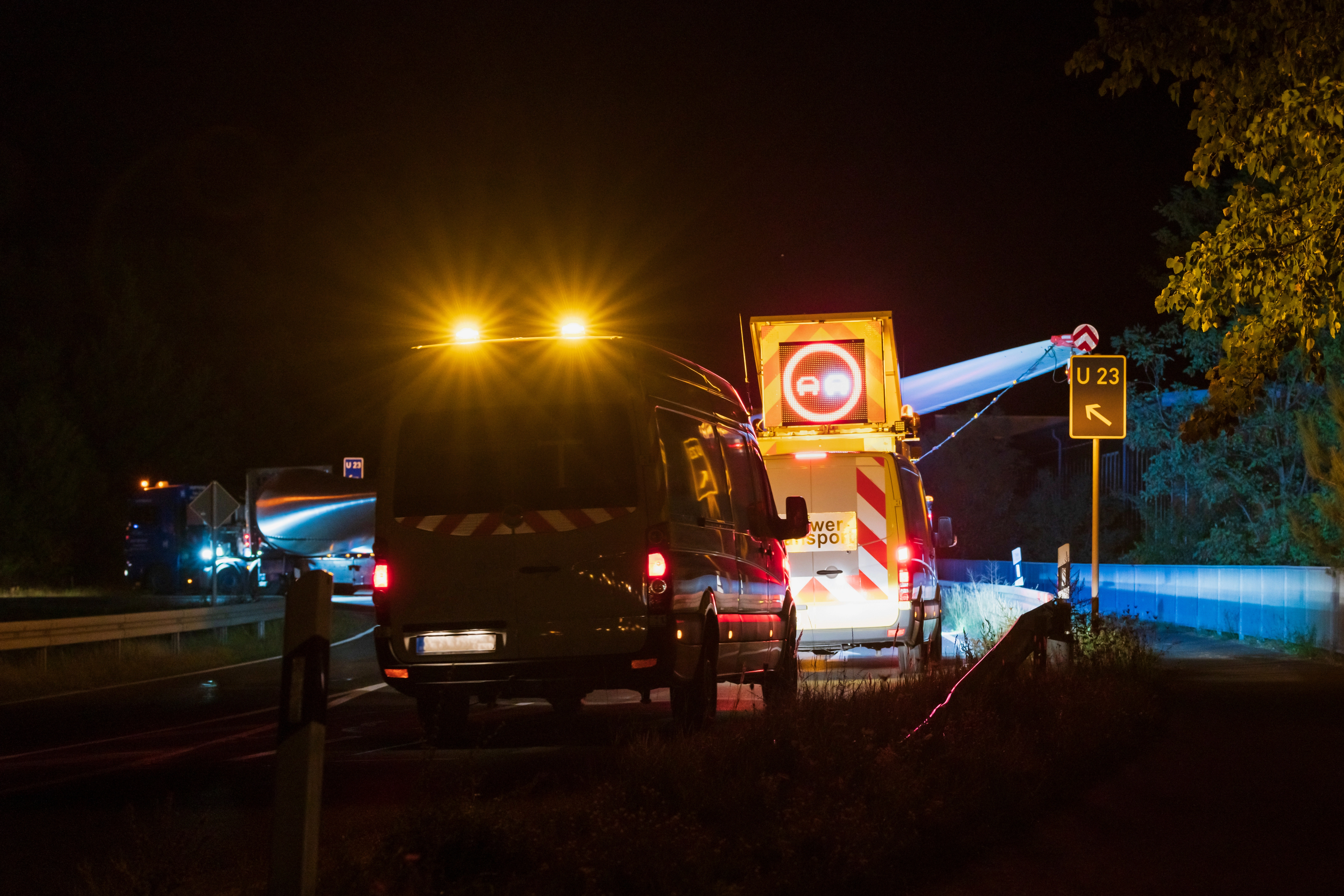 Schwertransport einer Windkraftkomponente bei Nacht mit Begleitfahrzeugen und Warnsignalen auf der Straße Schwertransport einer Windkraftkomponente bei Nacht mit Begleitfahrzeugen und Warnsignalen auf der Straße
