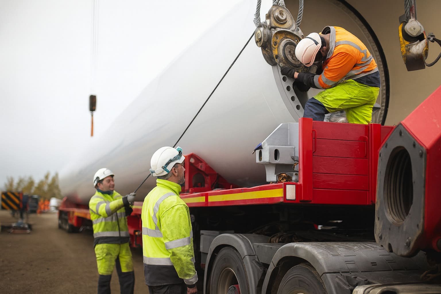 Arbeiter sichern ein Turmsegment einer Windkraftanlage auf einem Schwerlasttransporter mit Kranunterstützung – kontrollierte Gewichtsverteilung und präzise Planung Arbeiter sichern ein Turmsegment einer Windkraftanlage auf einem Schwerlasttransporter mit Kranunterstützung – kontrollierte Gewichtsverteilung und präzise Planung